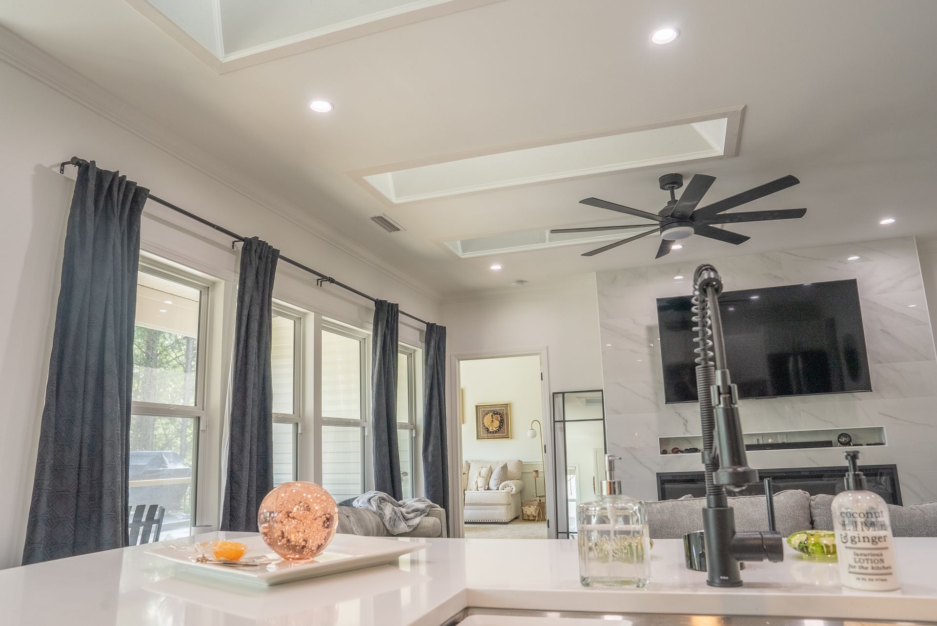 A kitchen island foregrounds an open-concept living area with dark curtains, a ceiling fan, and a wall-mounted television.
