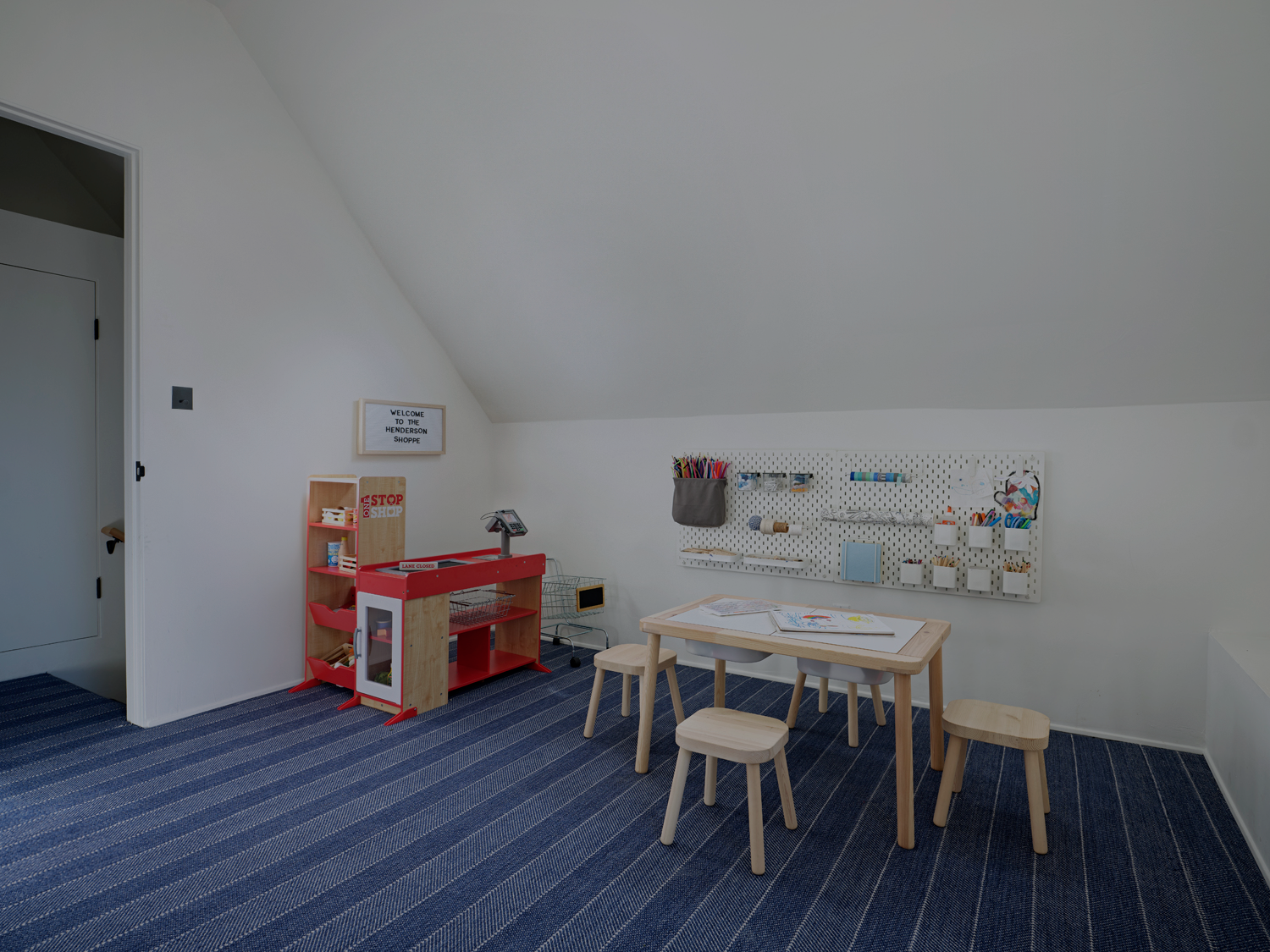 A child's playroom with a toy kitchen, a small wooden table with three stools, and a pegboard on a slanted white wall.