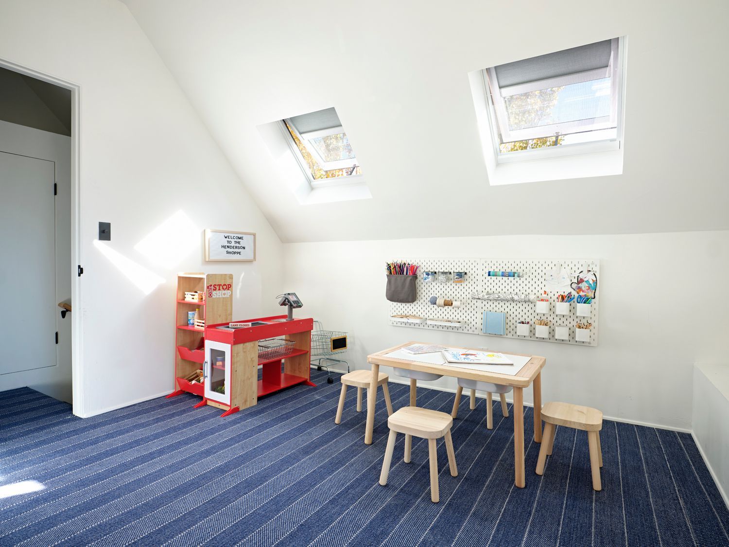 A child's play area with a red toy kitchen, a small wooden table, four stools, and a wall-mounted pegboard organizer.