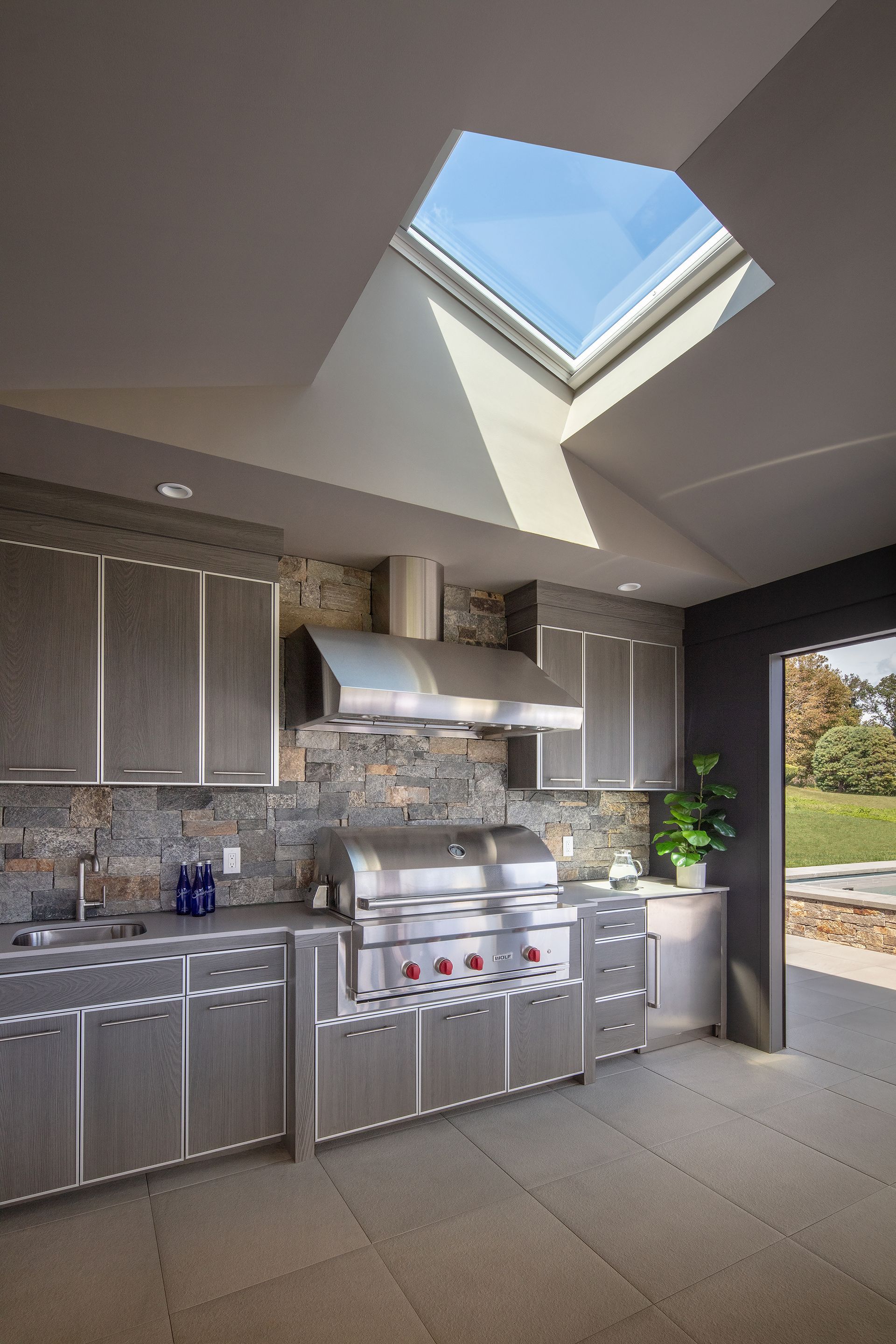 Modern outdoor kitchen with stainless steel grill, gray cabinets, stone backsplash, and a large skylight in the ceiling.