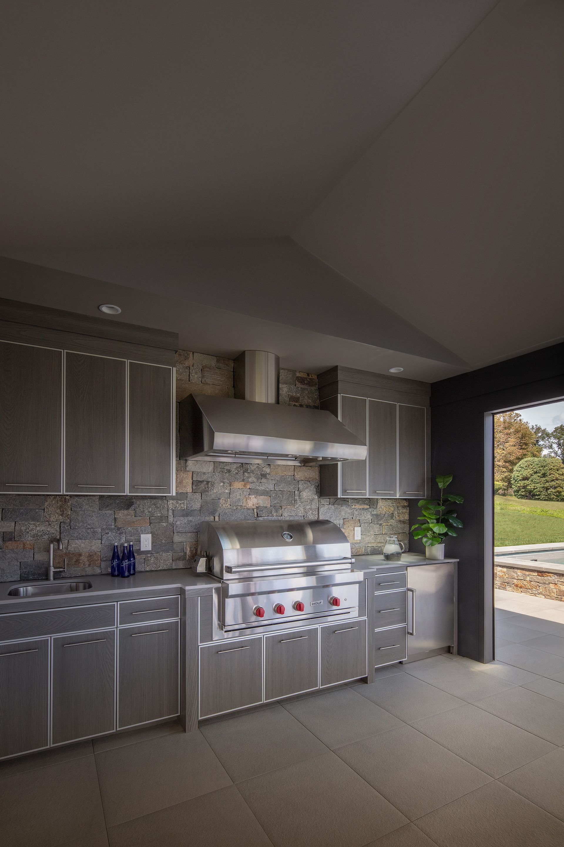 Modern outdoor kitchen with stone backsplash, stainless steel appliances, grey cabinetry, and an open entryway to a lawn.