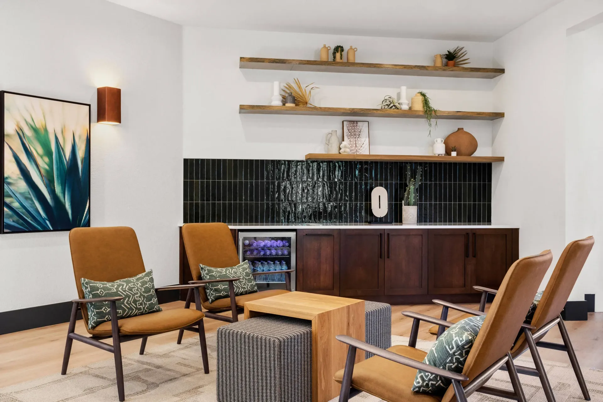 Communal lounge with tan mid-century chairs around a wooden coffee table and a dark tiled kitchenette at Briargate on Main apartments in Parker, CO.