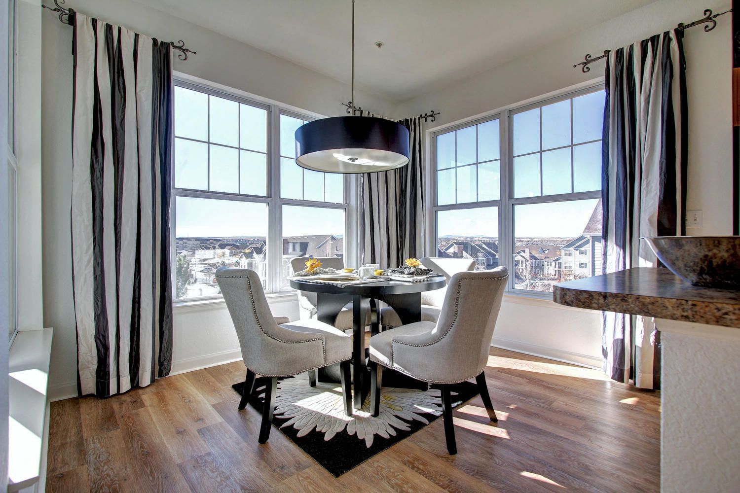 Round dining table with four upholstered chairs by large bay windows, sunlit at Briargate on Main apartments in Parker, CO.