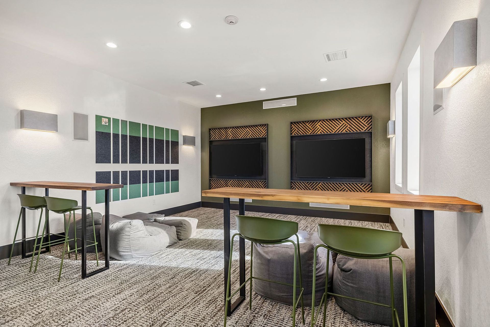 Indoor community lounge with two wall TVs, a long wooden counter, green stools, and bean bag seating at Briargate on Main apartments in Parker, CO.