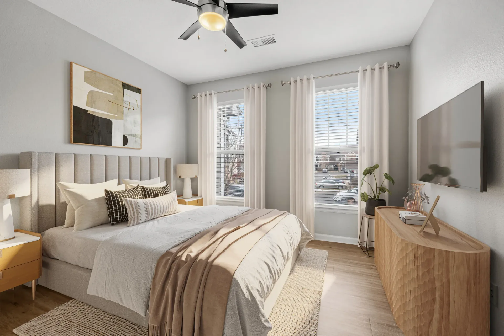 Bright bedroom with beige upholstered bed, two nightstands, dresser with TV, and large windows with white curtains at Briargate on Main apartments in Parker, CO.