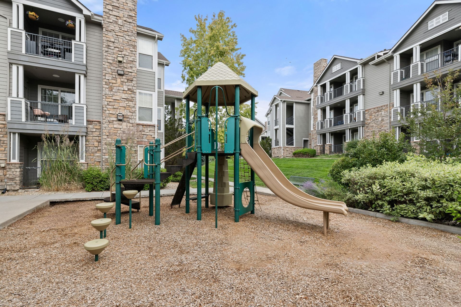 Apartment courtyard playground with green play structure, tan slide, and climbing steps on wood chips