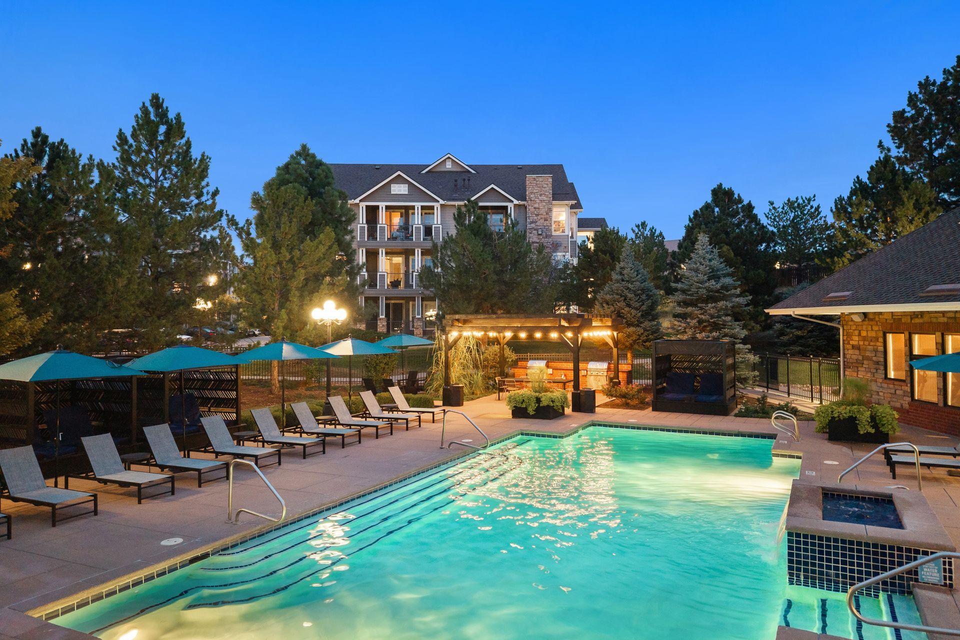 Resort pool at dusk with lounge chairs, lit cabana, and multi-story apartment building in the background