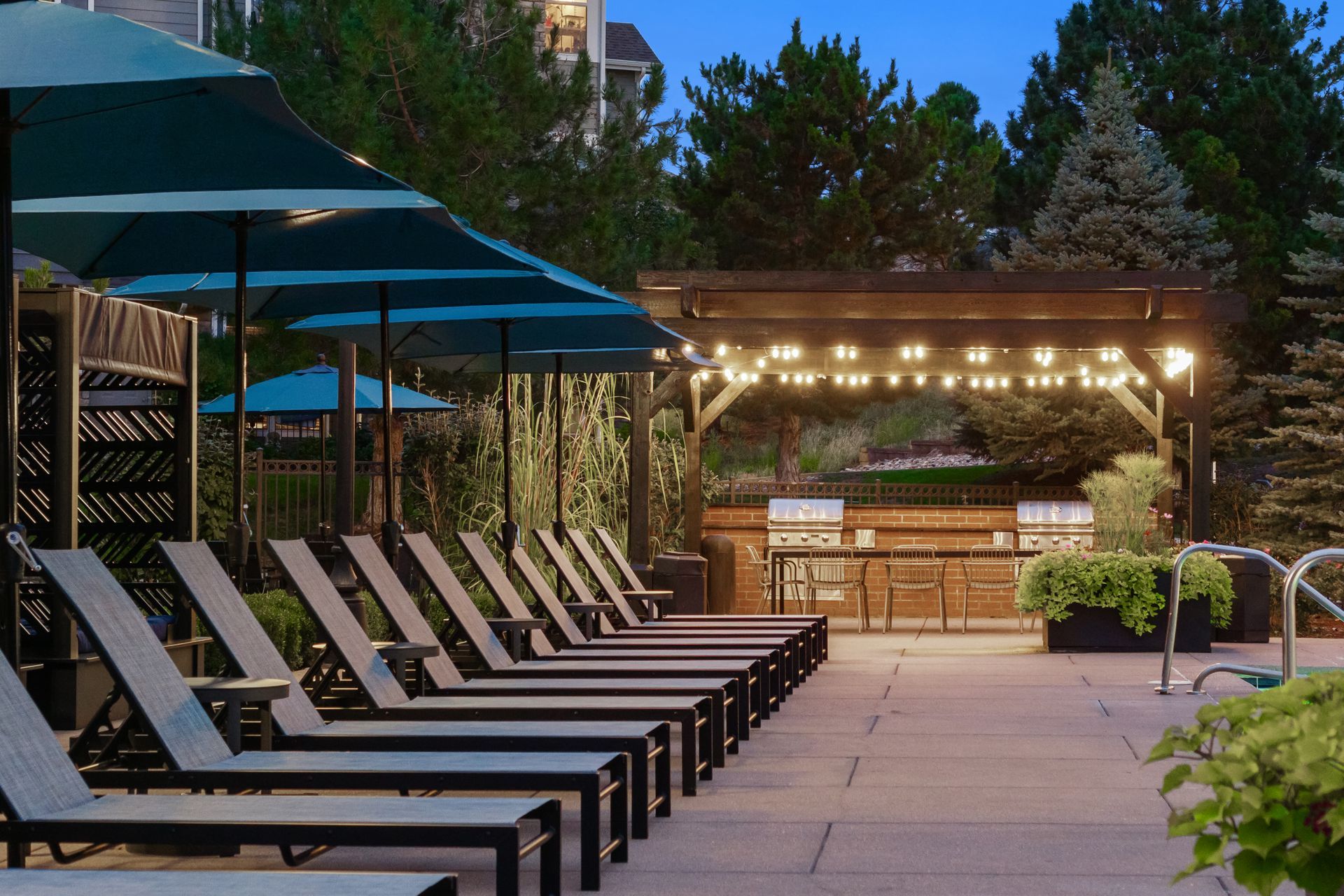 Poolside lounge chairs under blue umbrellas beside a lit outdoor bar at dusk