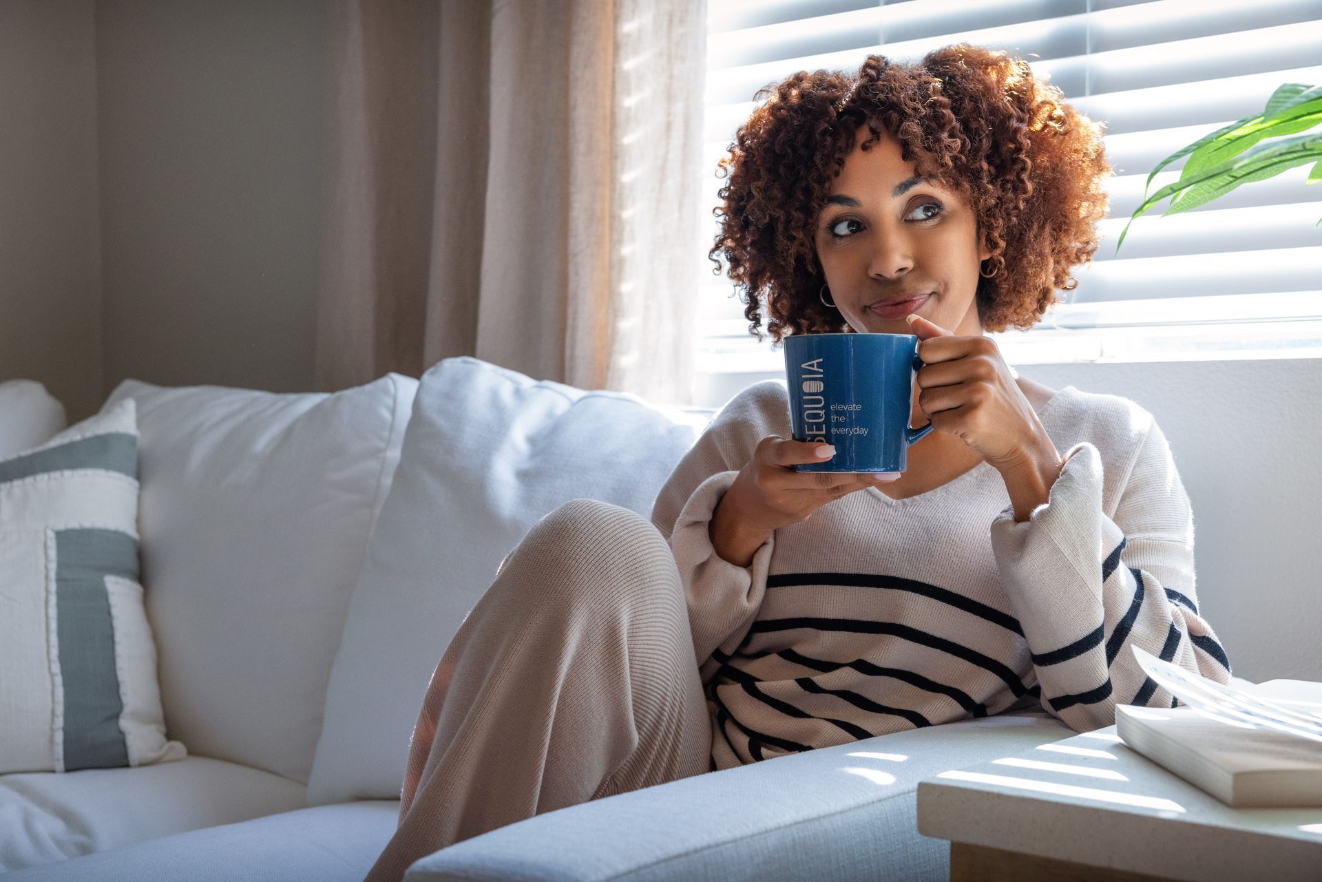 Woman on couch, holding blue mug, looking out window, sunlight streaming through blinds.