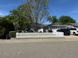 Suburban house with white picket fence, leafless tree, and parked cars under a clear blue sky