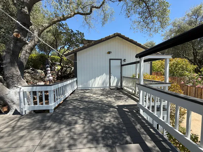 Wooden deck walkway leading to a white house amid trees, with railings and dappled sunlight.