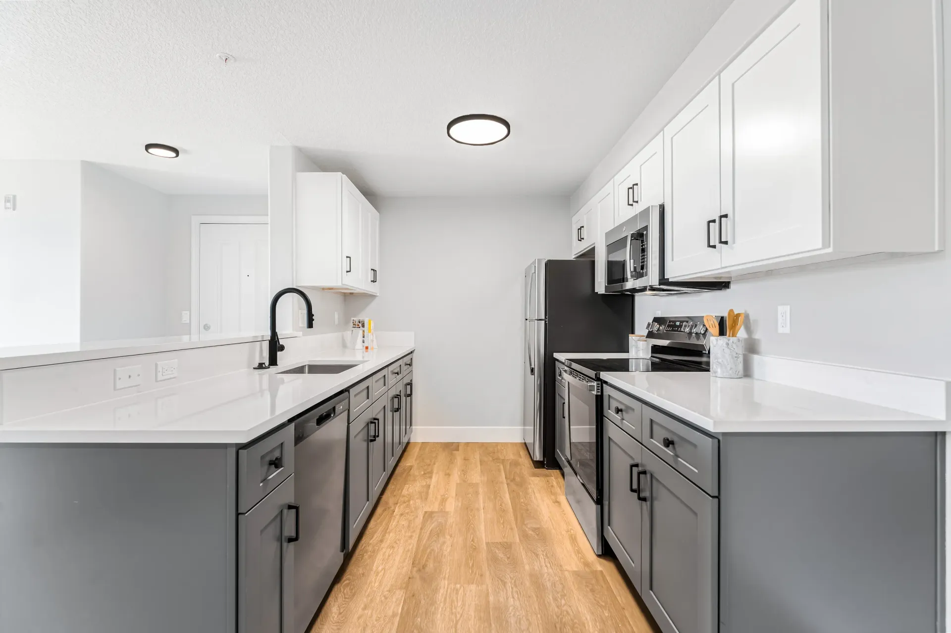 Modern apartment kitchen with white upper cabinets, gray lower cabinets, stainless steel appliances, and wood-look flooring.