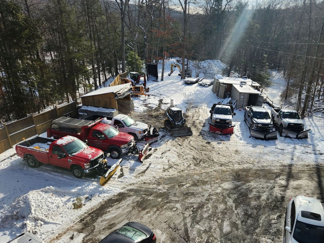 Snow-covered parking lot with several parked cars and trees in the background.
