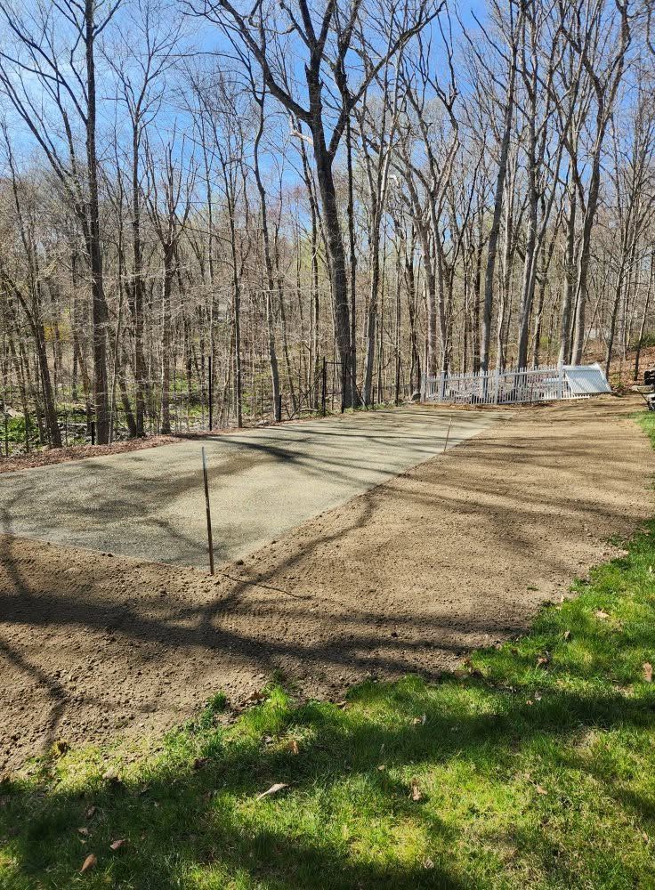 Concrete slab in a wooded yard with bare trees and grass under a clear blue sky.