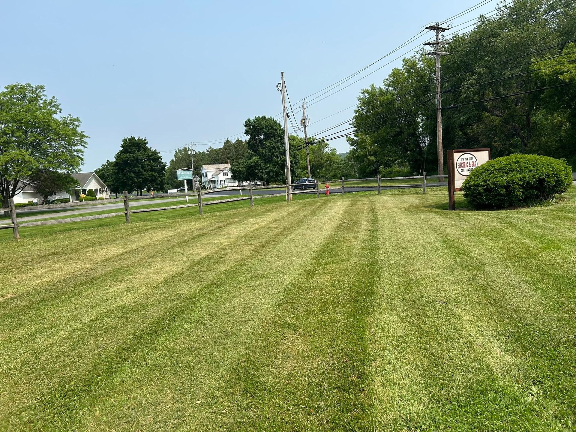 Mowed grassy field beside a road, with trees, utility poles, and a small sign under a clear blue sky.