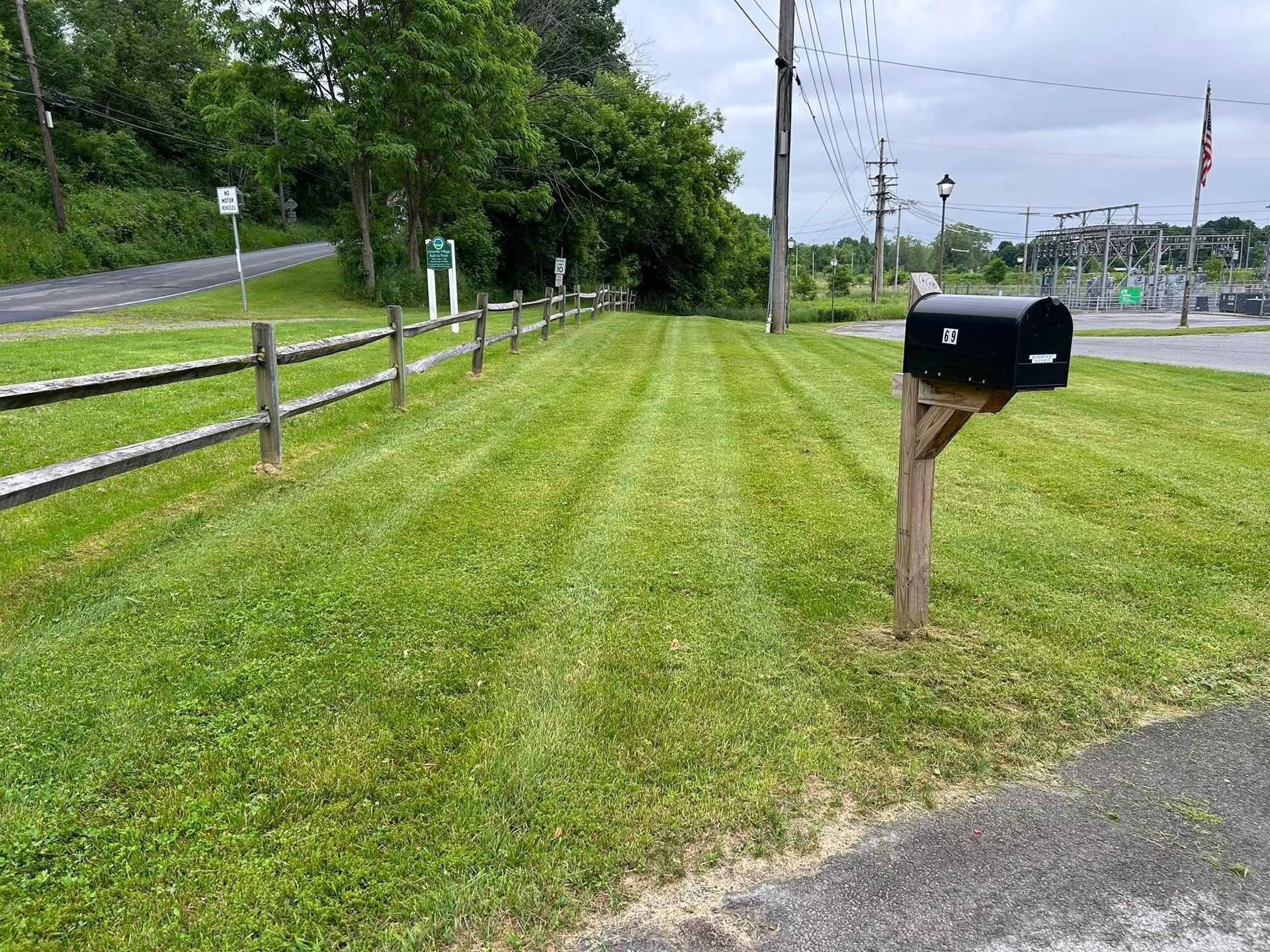 Grassy roadside with a wooden mailbox post and black mailbox beside a fenced path under cloudy skies