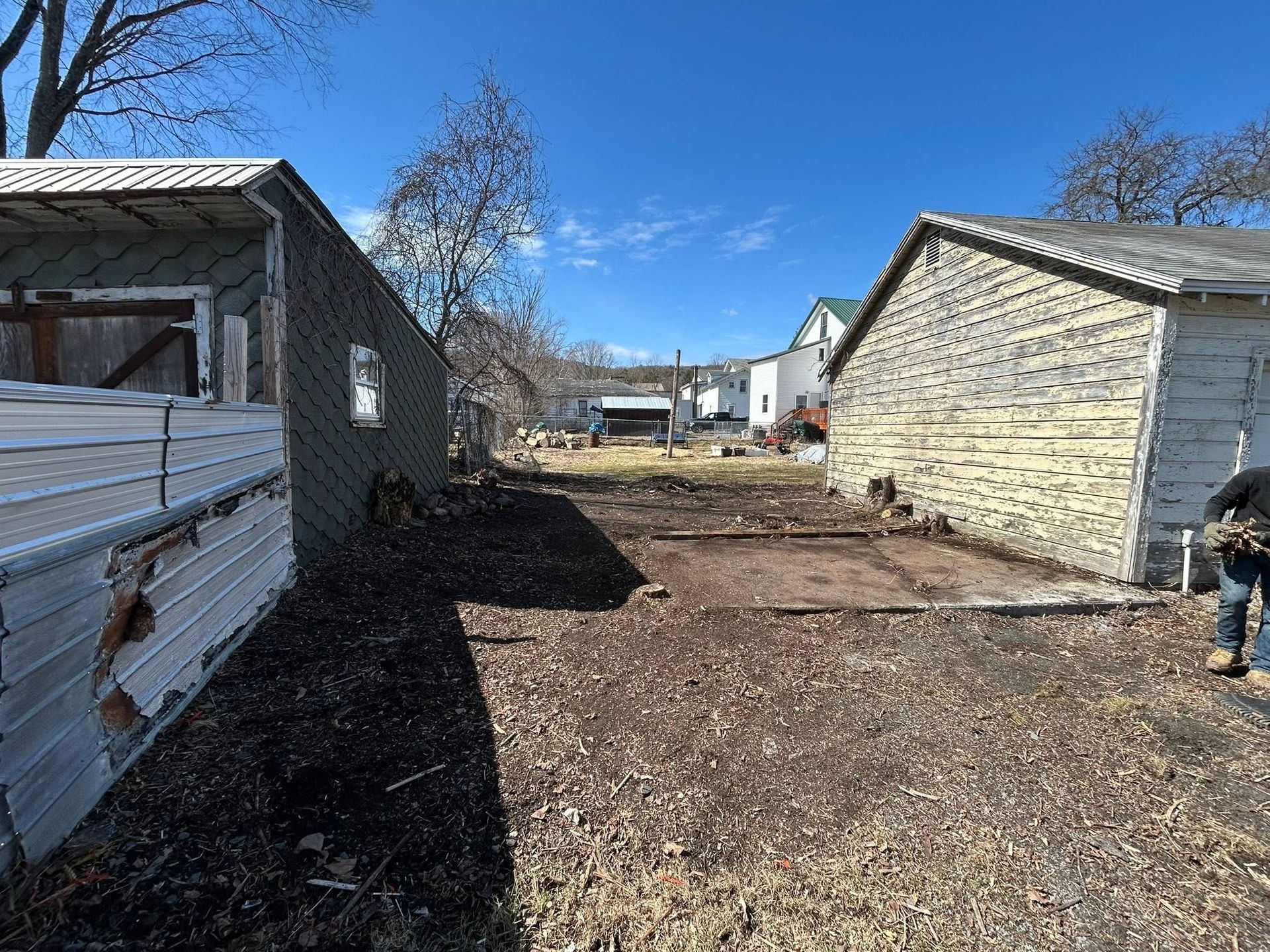 Narrow dirt path between two buildings with a person standing at the right edge under a blue sky.