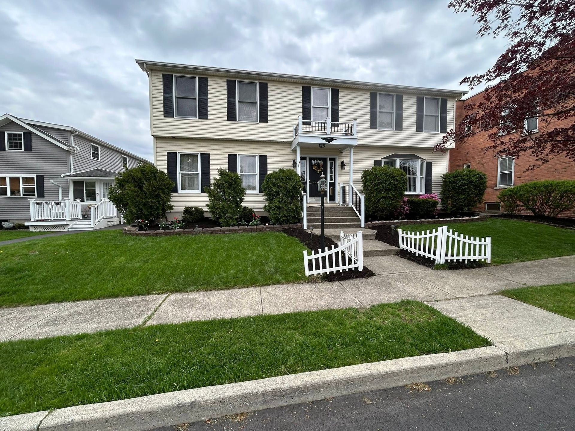 Two-story cream house with dark shutters, white trim, and a small front lawn and walkway in a suburban neighborhood