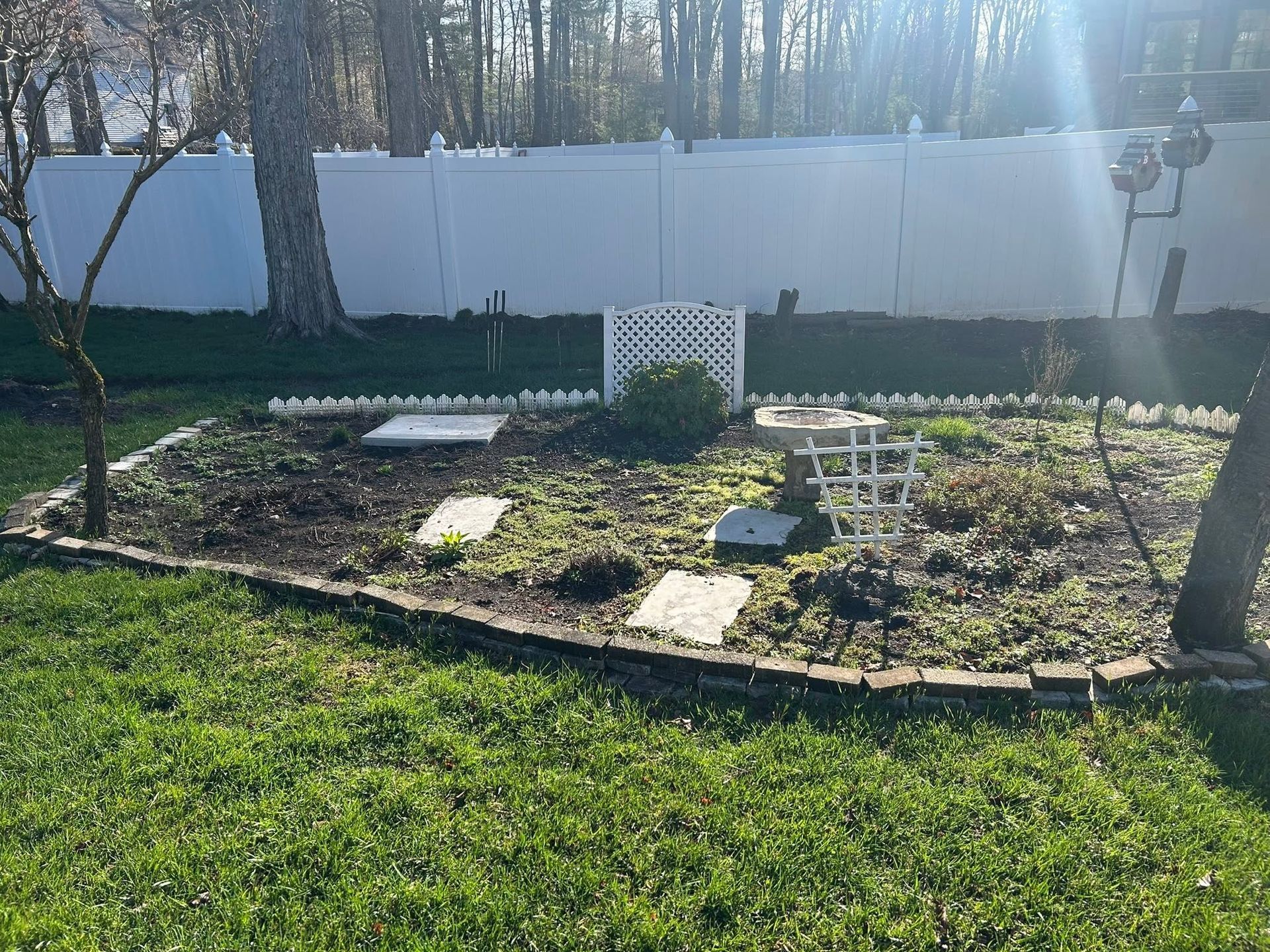 Sunlit backyard garden bed with stepping stones, white fence, and trees in the background
