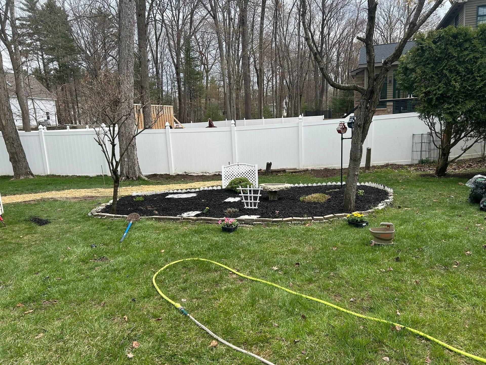 Backyard garden bed with white fence, lawn, and hose in foreground