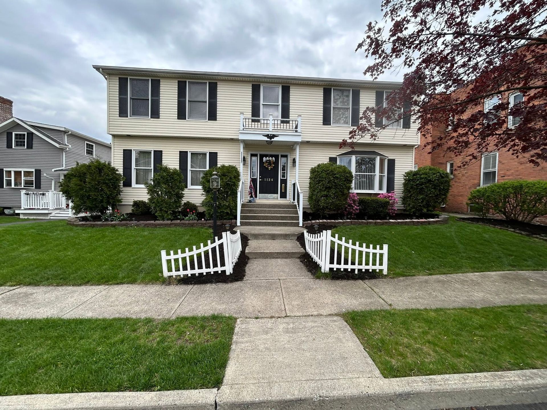 Two-story cream house with black shutters, front steps, white picket fence, and a green lawn.