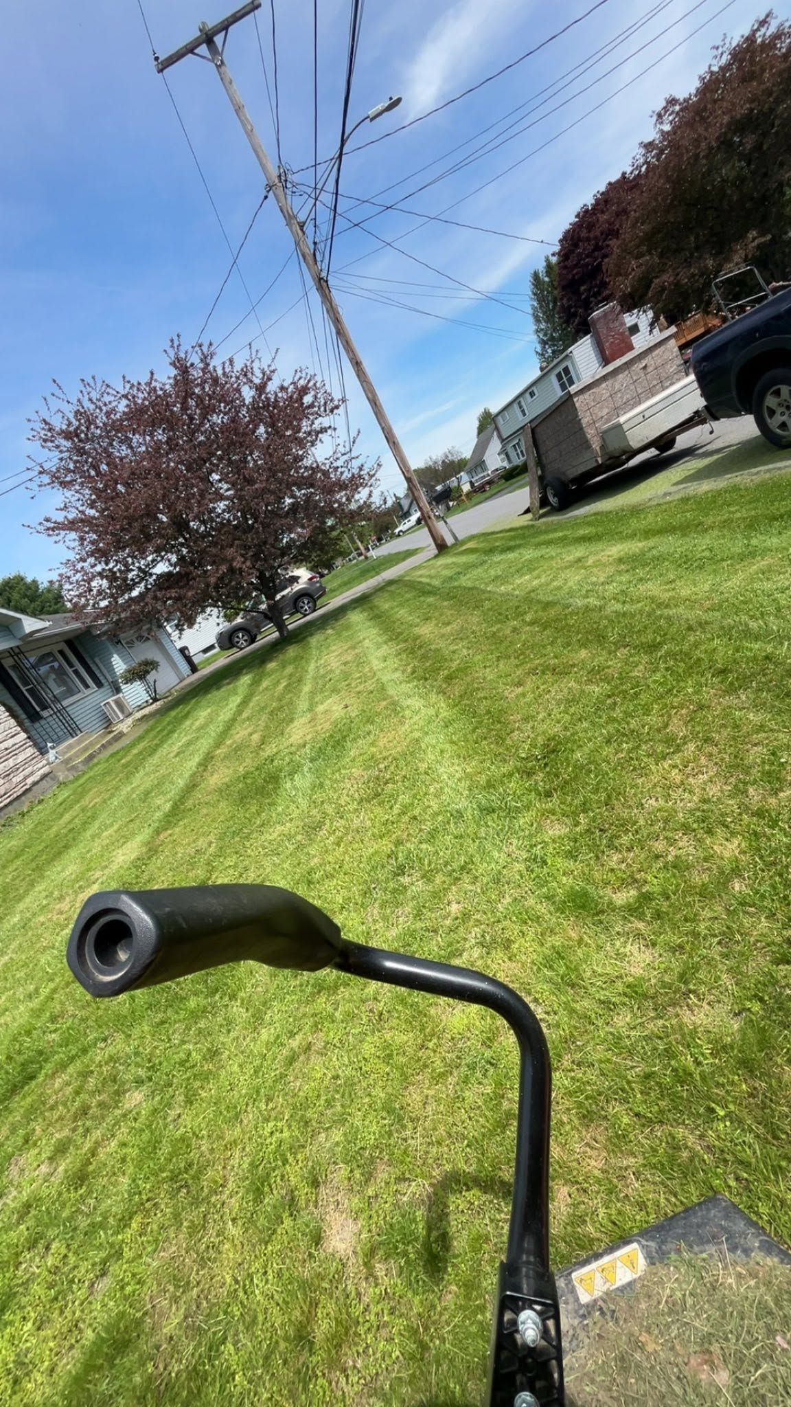 Tilted view of a grassy roadside with utility lines, parked vehicles, and a black scooter handlebar in the foreground