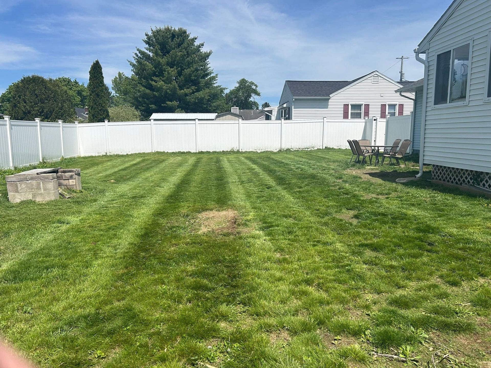Backyard with green lawn, white fence, and a house on the right under a blue sky