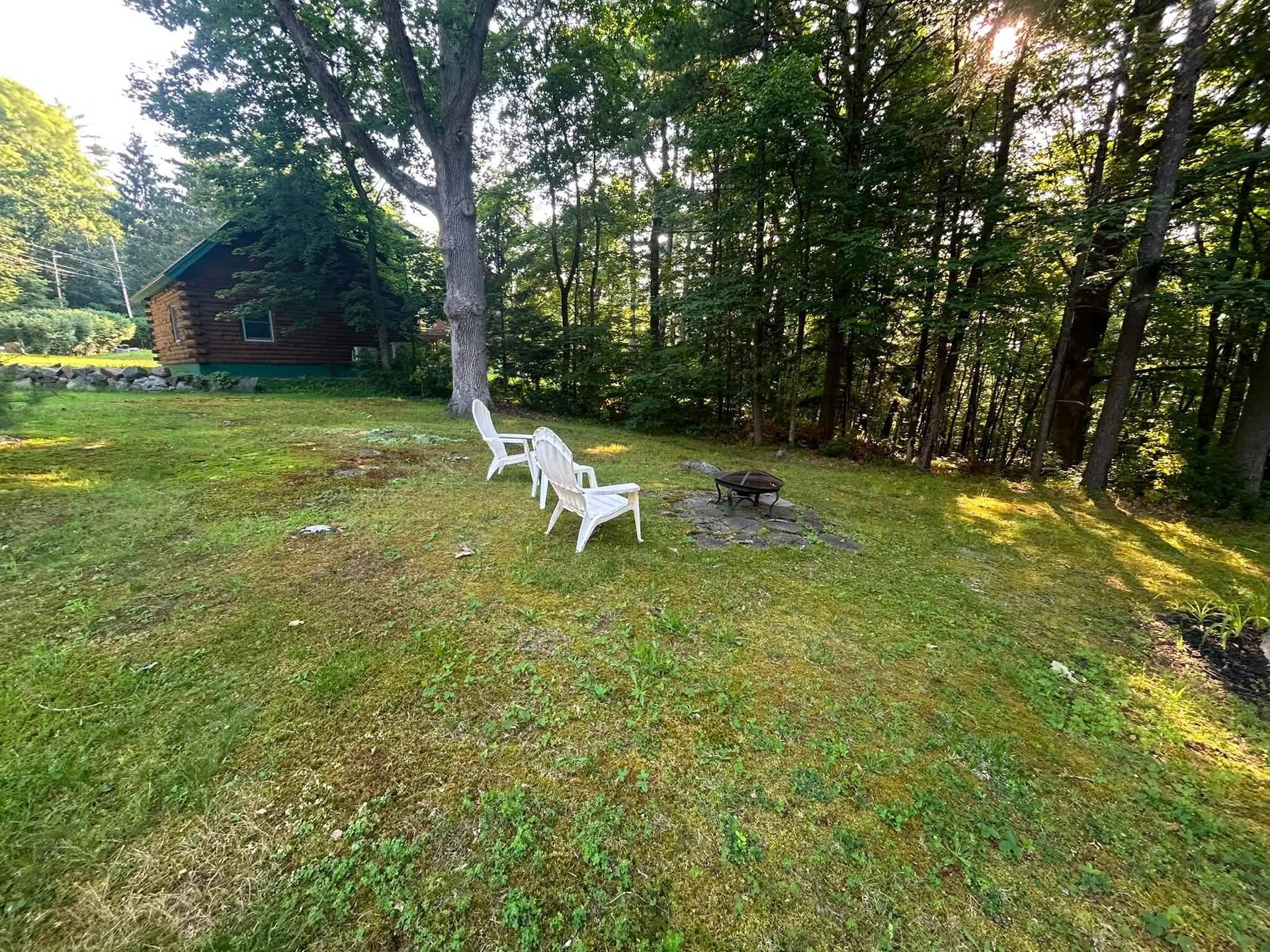 Two white chairs by a fire pit on a grassy yard beside a wooded cabin at sunset