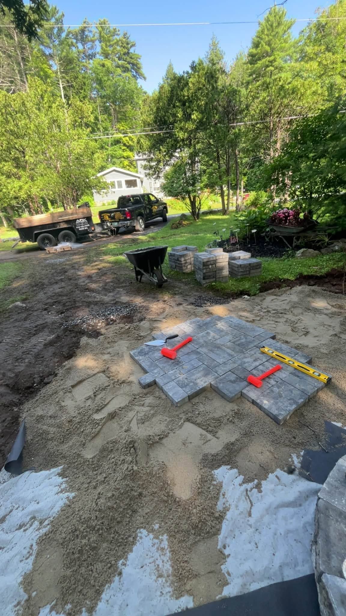Construction site with wheelbarrow, red tools, and concrete pavers set in muddy ground near trees.