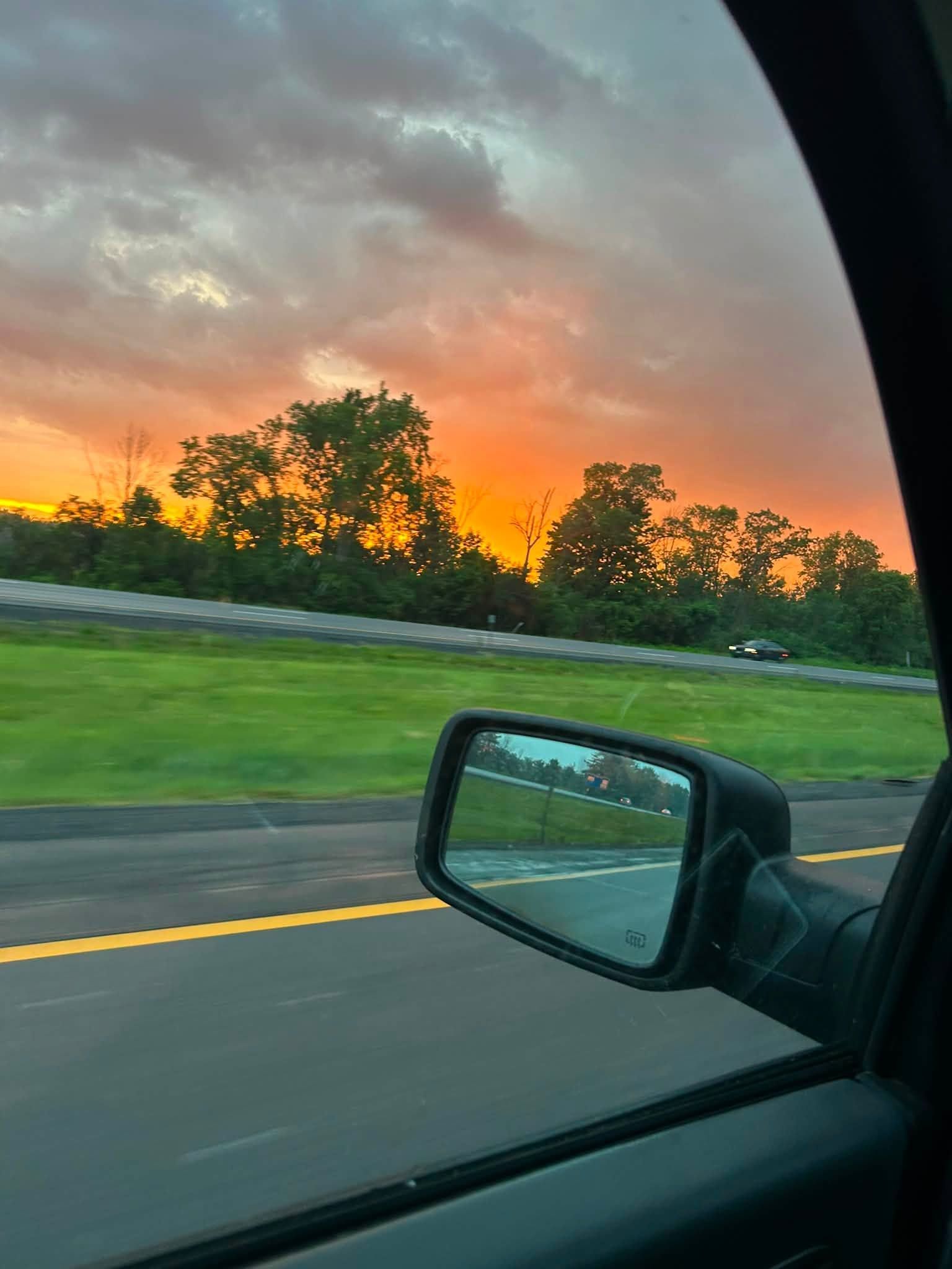 Sunset over a highway, seen from a car window with side mirror and trees silhouetted against orange sky