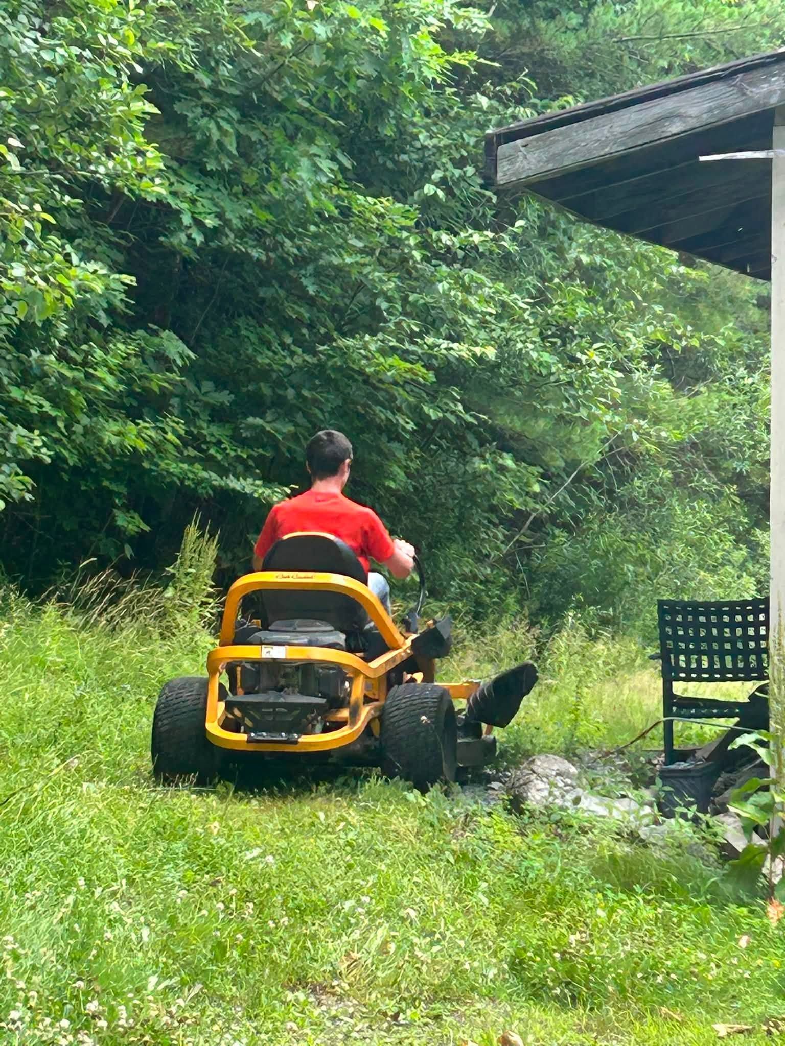 Person mowing grass on a yellow riding mower beside a house, surrounded by trees.