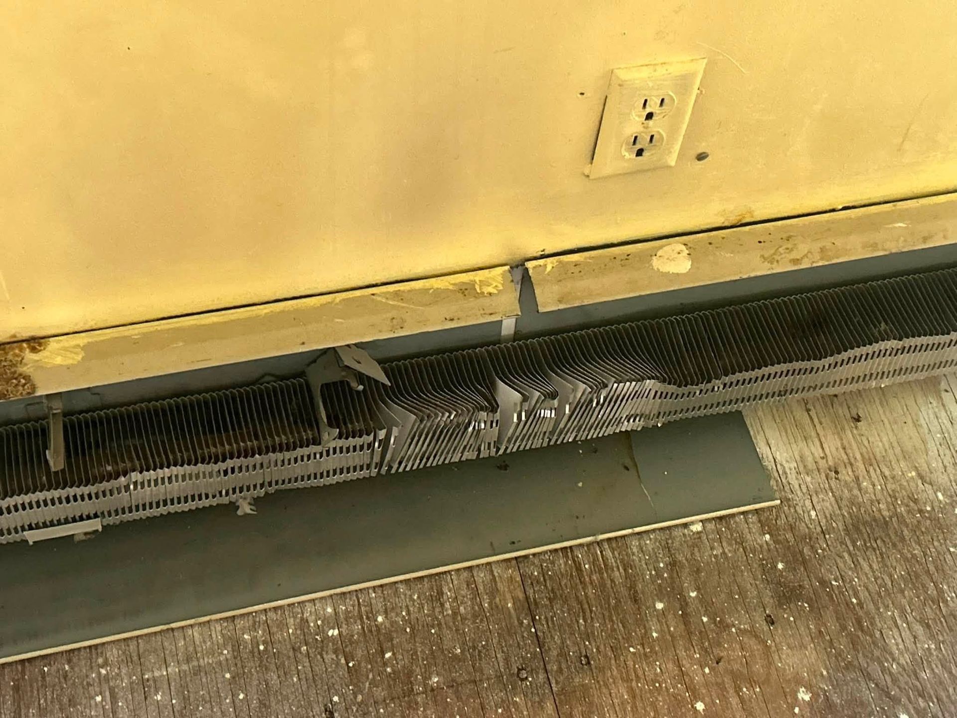 Damaged baseboard heater below a wall outlet, with a bent vent cover and debris on the floor
