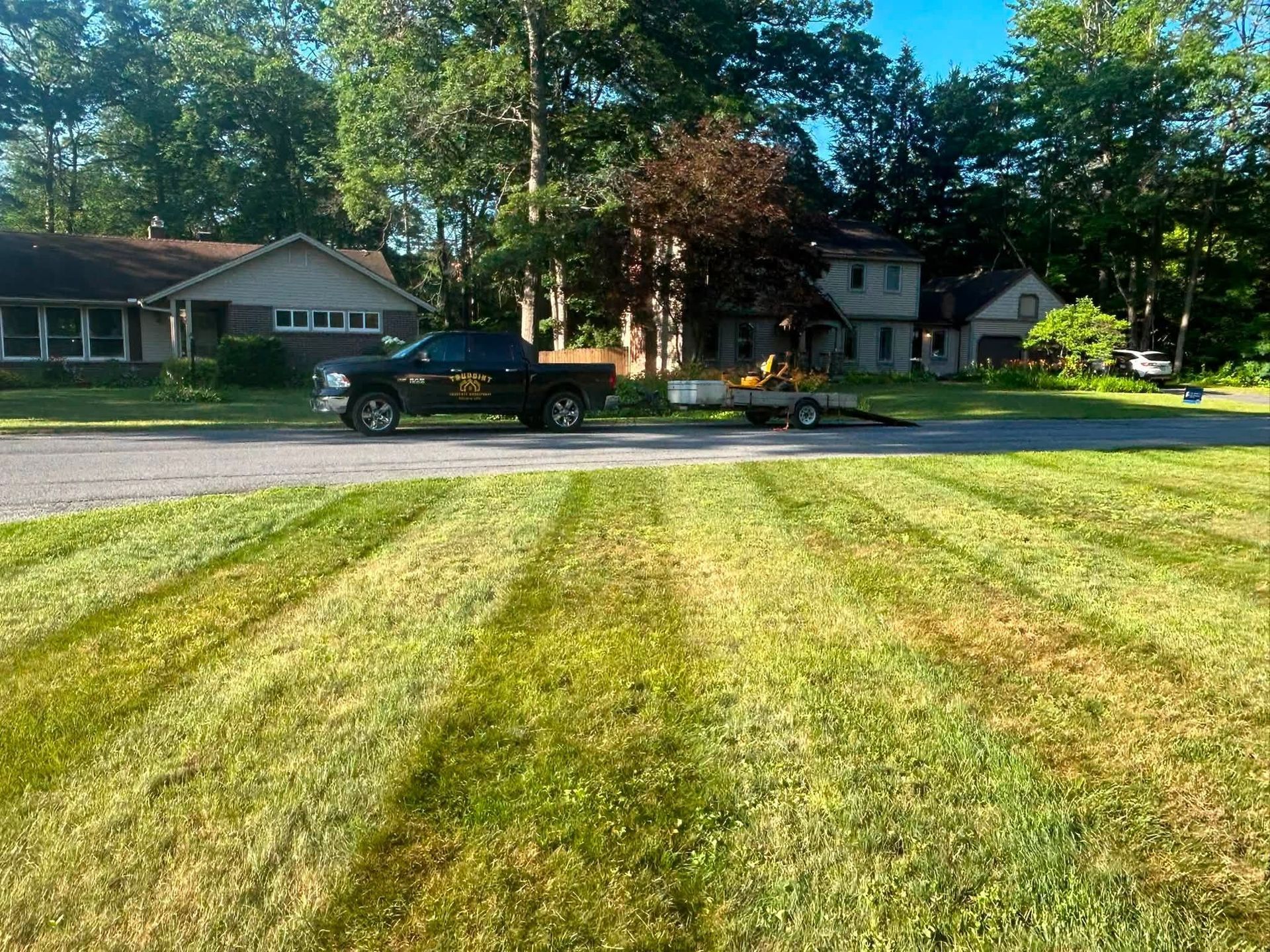 Freshly mowed lawn with a pickup truck towing a trailer on a suburban street, houses and trees behind.