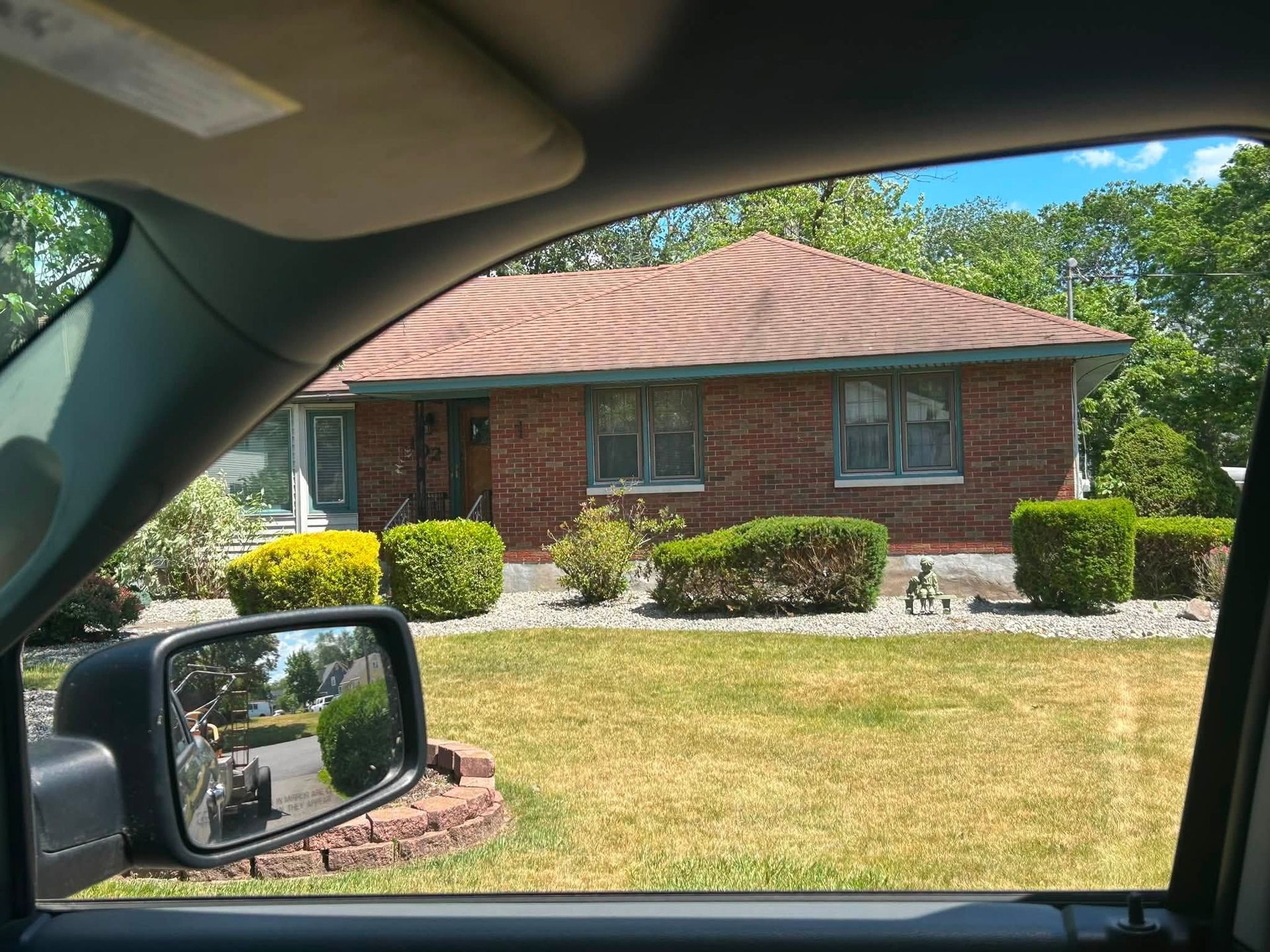 Brick house with red roof and trimmed bushes seen from inside a parked car on a sunny day