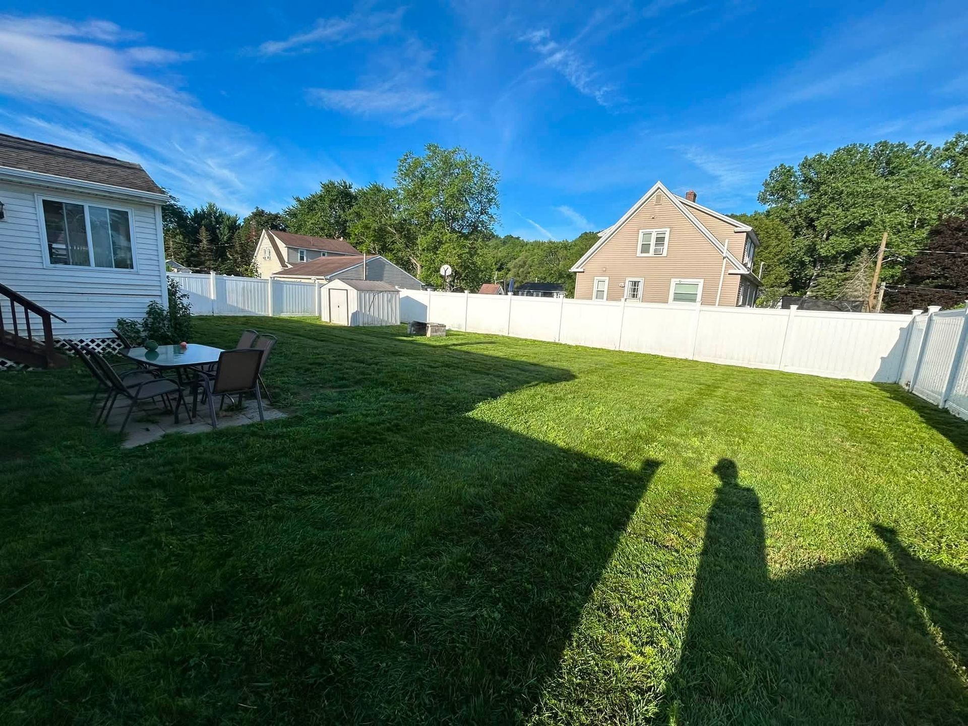 Backyard with green lawn, white fence, patio chairs, and a house under a blue sky.