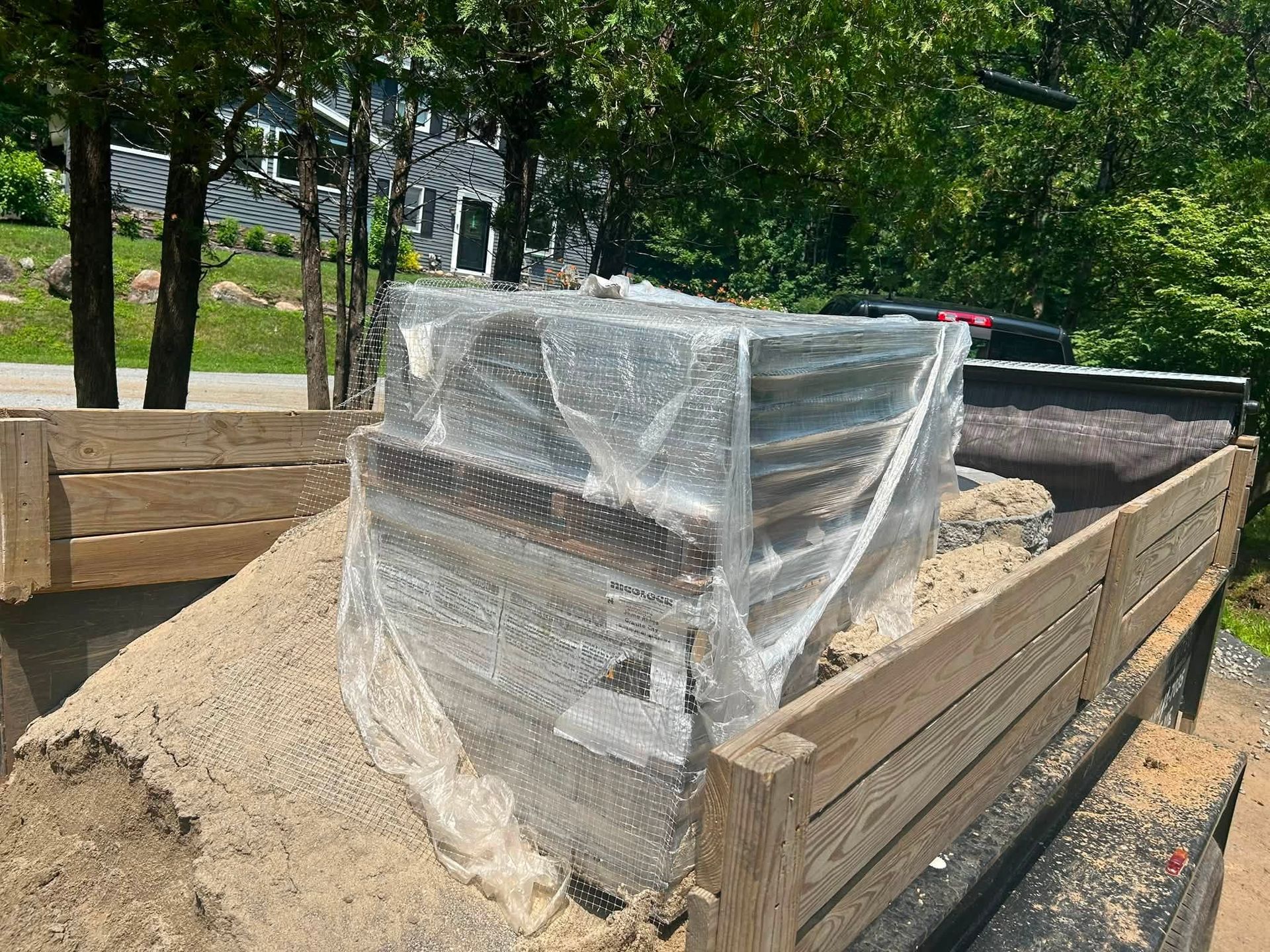 Pallets of sand on a flatbed trailer, wrapped in plastic, outdoors by trees