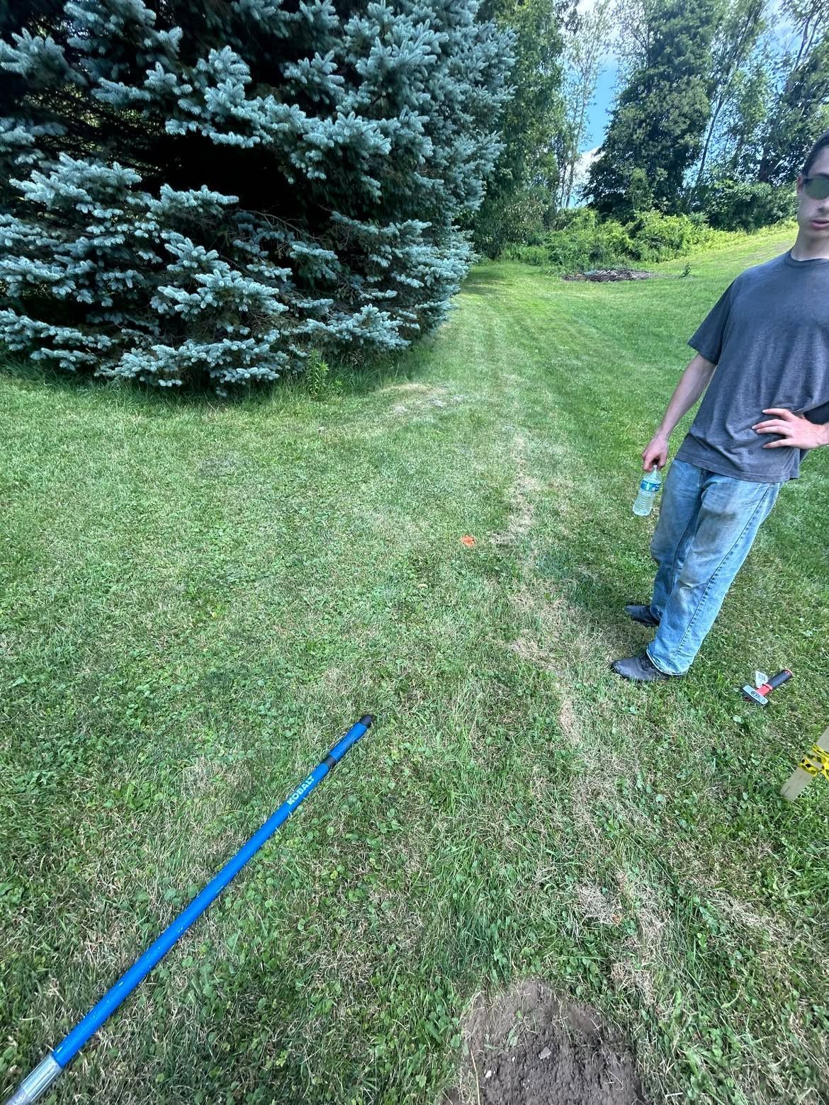 Man standing on a grassy lawn near evergreen trees, holding a blue bottle and a blue pole in the foreground.