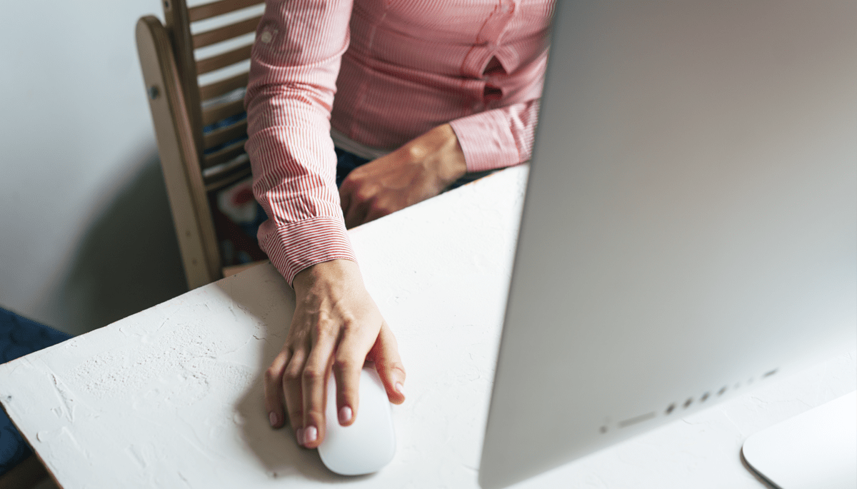 Pessoa usando um computador, mão no mouse, camisa rosa, mesa branca.