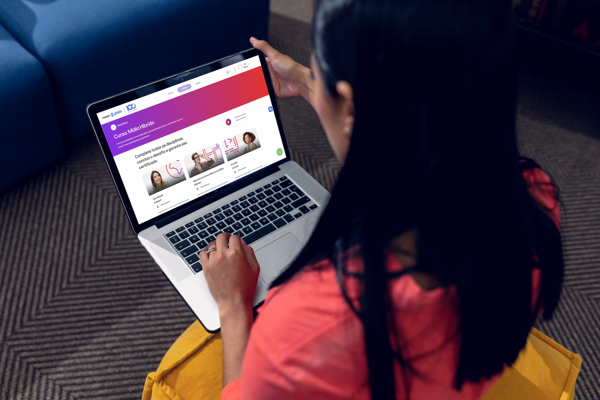 Pessoa visualizando conteúdo online em um laptop; sentada em uma almofada amarela, com tela roxa e vermelha.