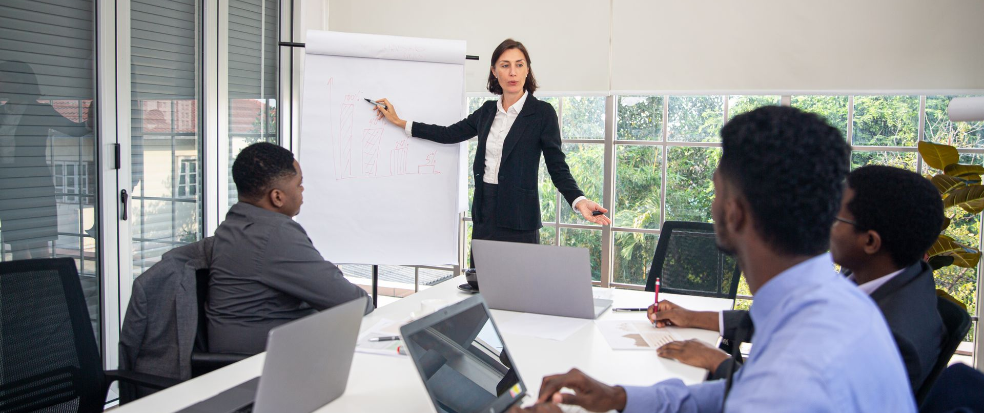 Mulher de negócios fazendo uma apresentação para colegas em uma sala de conferências. Ela aponta para um flip chart.