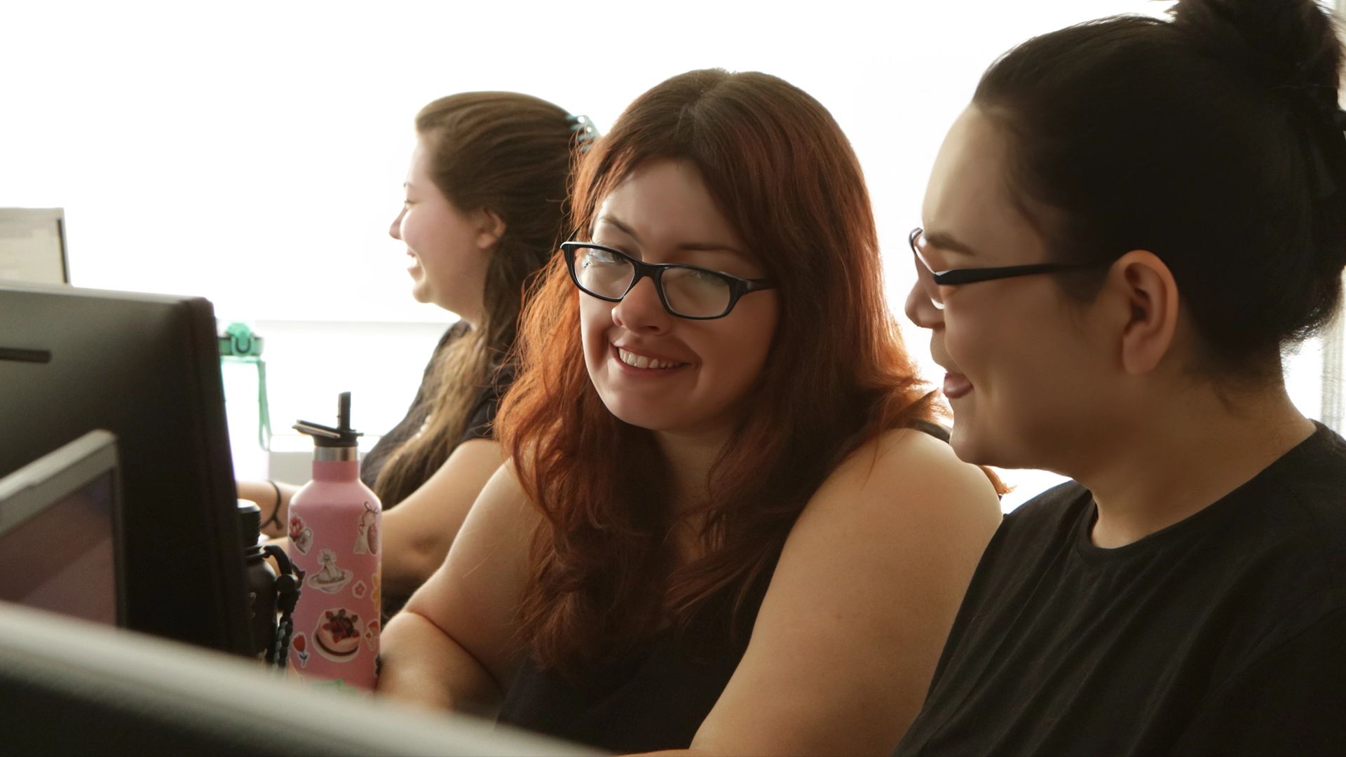 Três pessoas trabalhando juntas em mesas; duas delas estão sorrindo e olhando para a tela do computador.