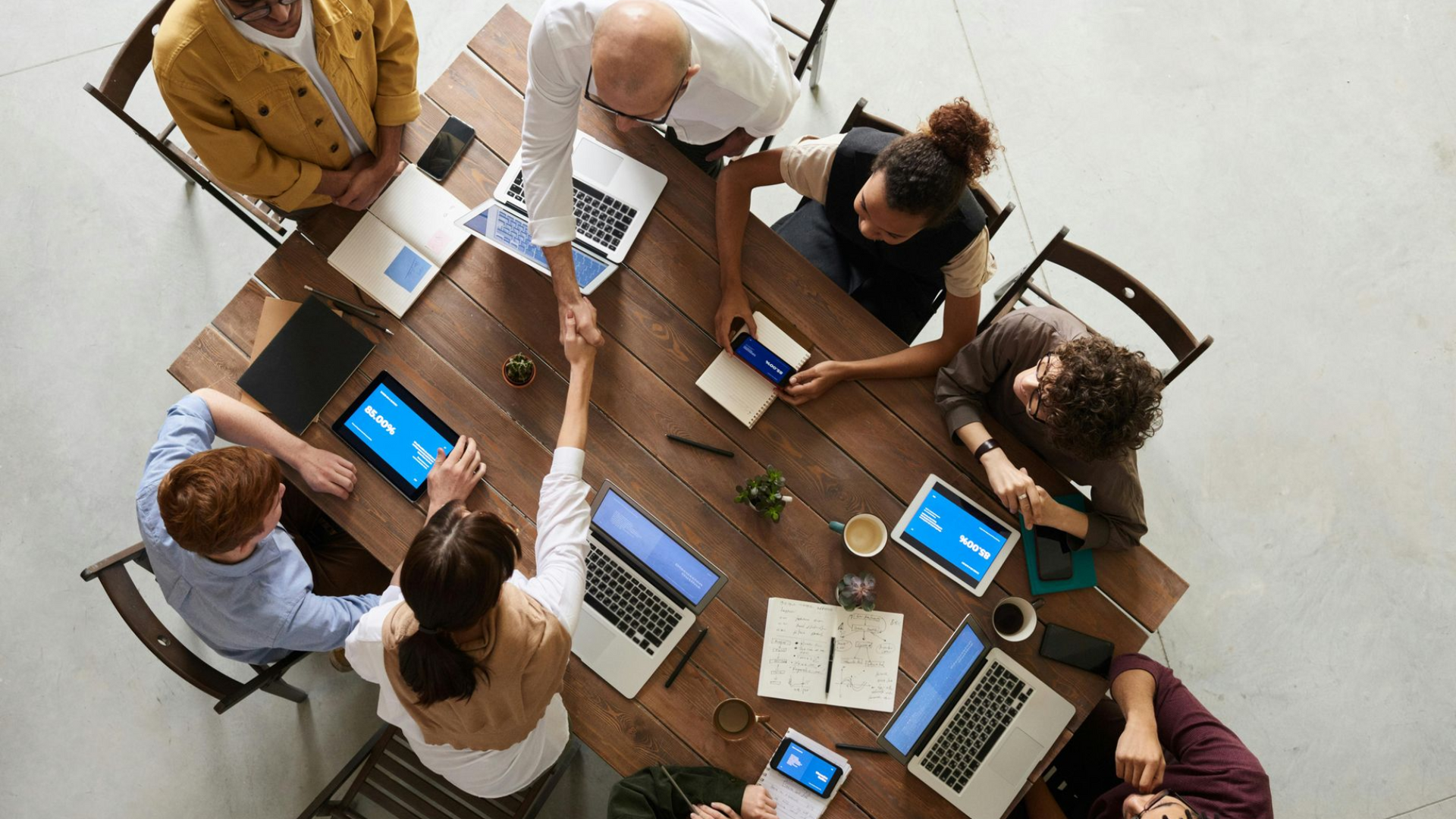 Pessoas apertando as mãos em volta de uma mesa de madeira com laptops e tablets.