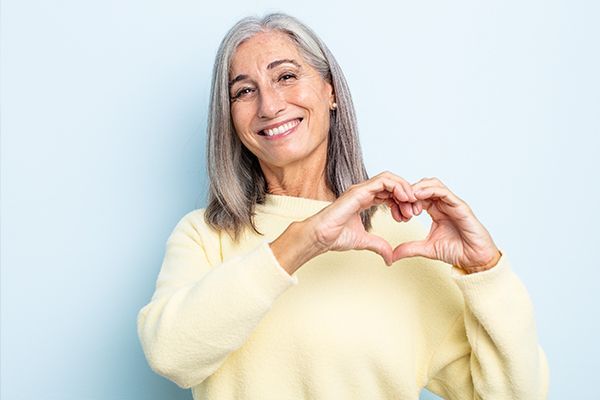 Smiling woman in a pale yellow sweater forming a heart with her hands against a light blue background