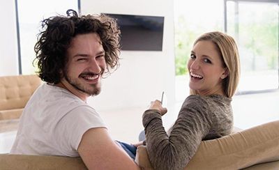 Smiling couple on a beige couch in a bright living room, looking back toward the camera.