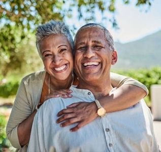 Two smiling people embracing outdoors in sunlight, with trees and hills in the background