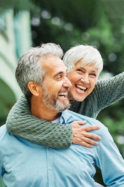 Two smiling adults outdoors, one hugging the other from behind