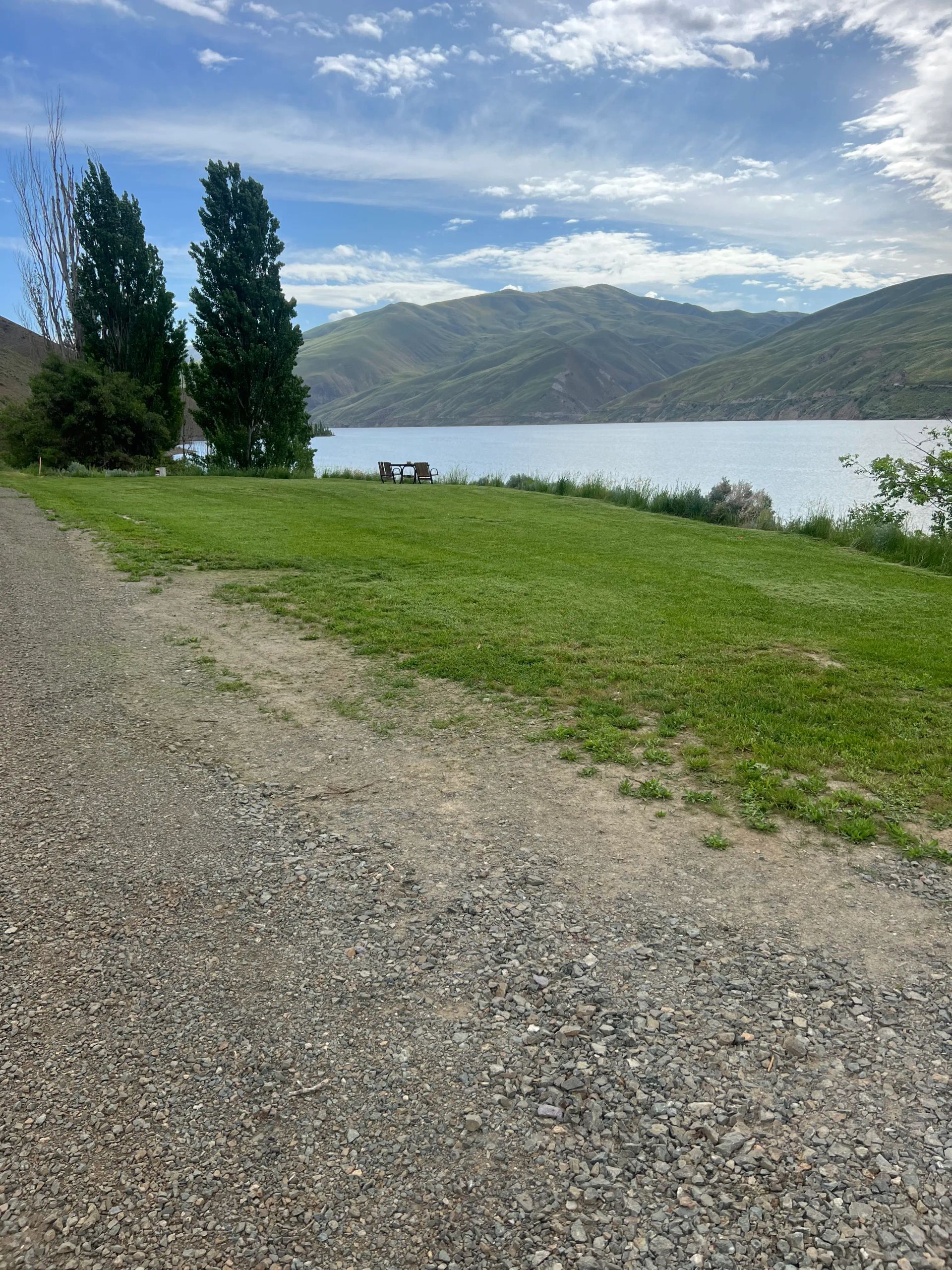 Gravel path beside a grassy lakeshore with tall trees and mountains under a cloudy sky