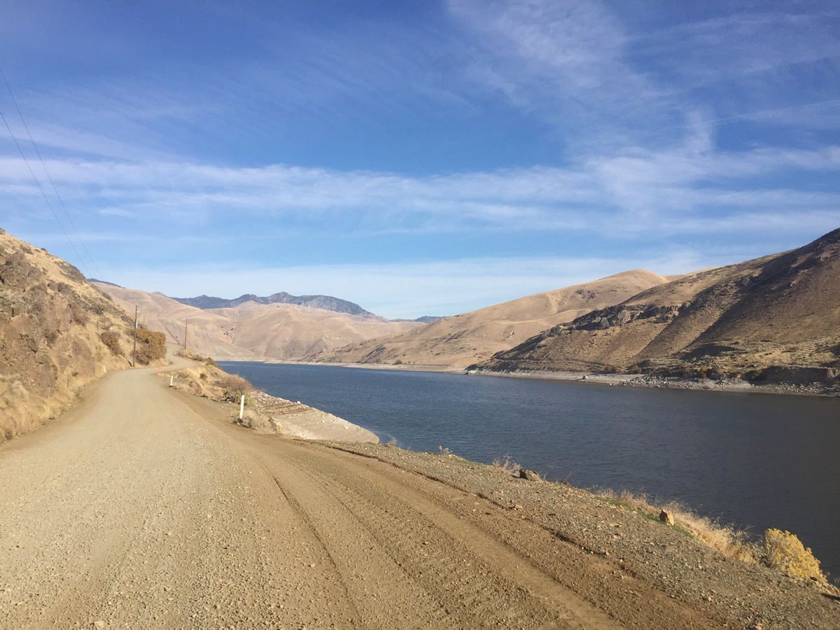 Desert road beside a calm blue river or lake, with rocky hills and clear sky