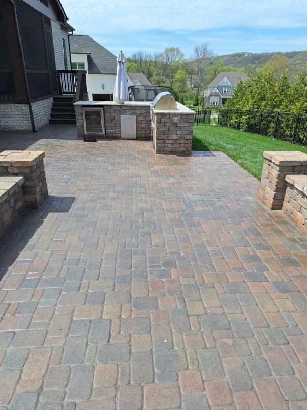 Brick patio and walkway beside a house, with stone planters and a grassy yard overlooking trees and hills.