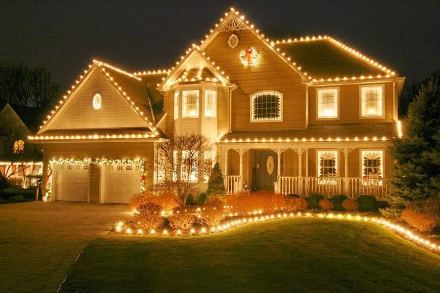 Two-story house at night outlined with warm white holiday lights, glowing in a decorated front yard.